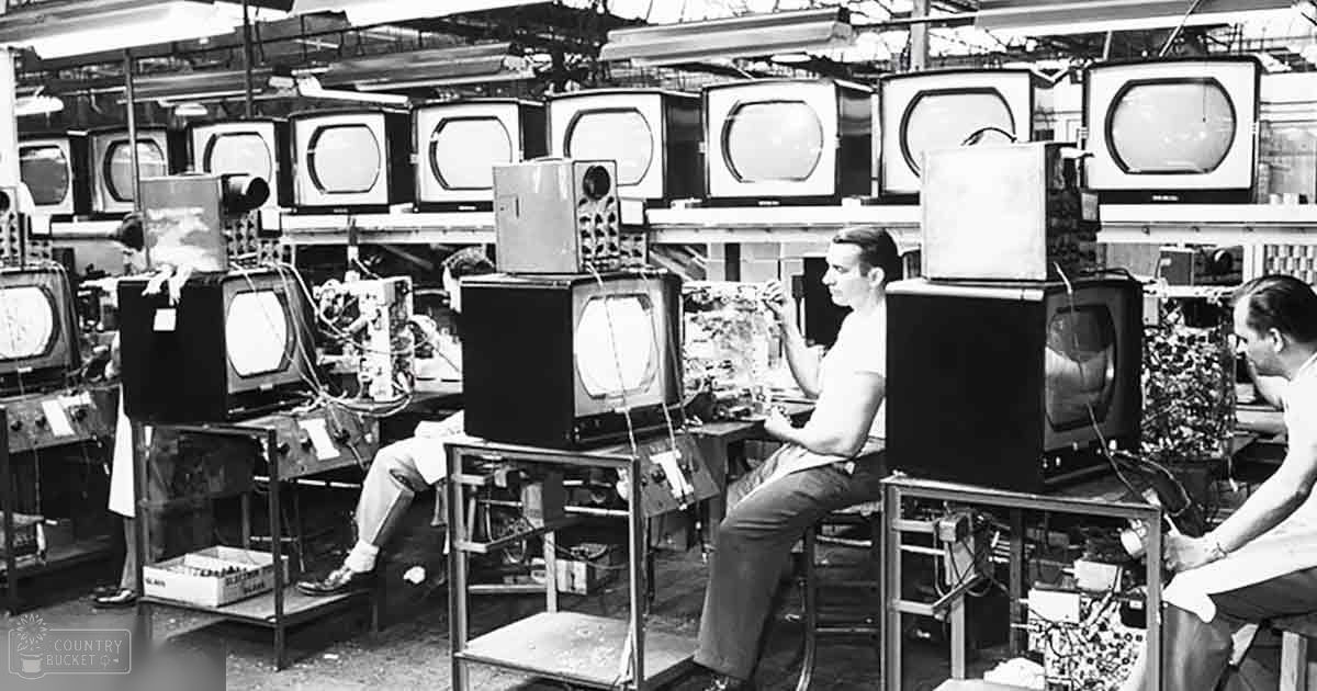 Adult males wearing white t-shirts work on television sets in a factory-like setting filled with rows of assembled or tested televisions under artificial lighting.