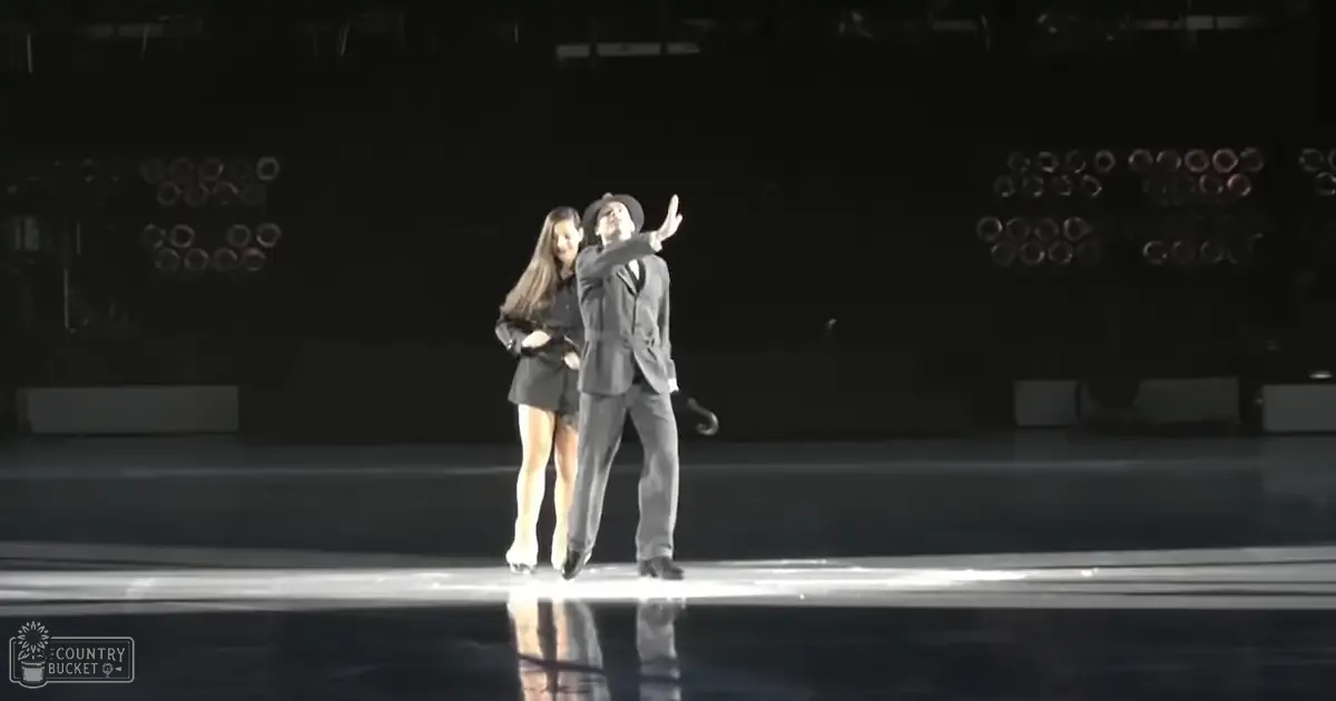 A couple performing on an indoor ice rink during a skating event; male in a black suit with white shirt, female in a dark dress or skirt.