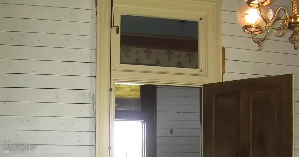 A residential interior with a partially open wooden door, white shiplap walls, decorative transom window, and a hanging chandelier emitting natural light.