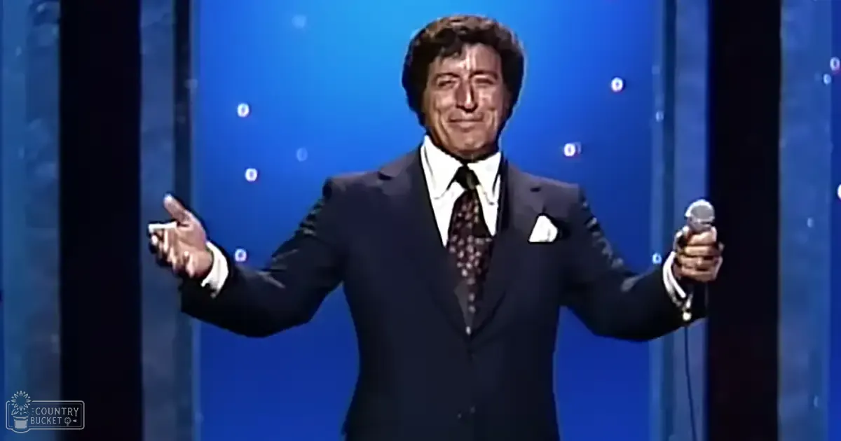 A middle-aged male performer in a dark suit with a patterned tie, holding a microphone on an indoor stage or studio set up for performance.
