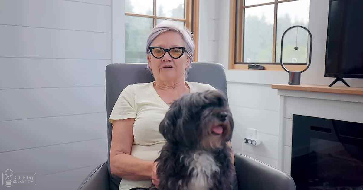 An older adult female in a living room with a small Shih Tzu or similar dog. The setting includes a dark gray armchair, white paneling on the wall, and natural daylight illuminating the space.