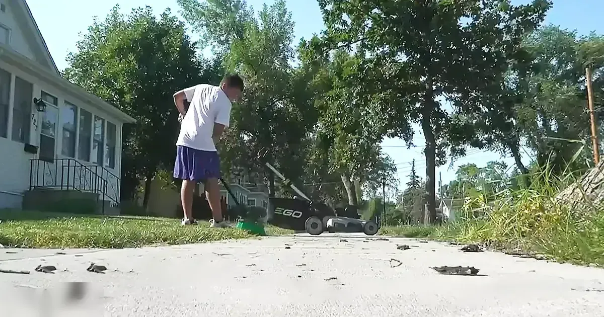 A young adult male wearing a white short-sleeved crew neck T-shirt, purple cuffed shorts, and white sneakers with dark accents mowing grass in front of a house on a sunny day.