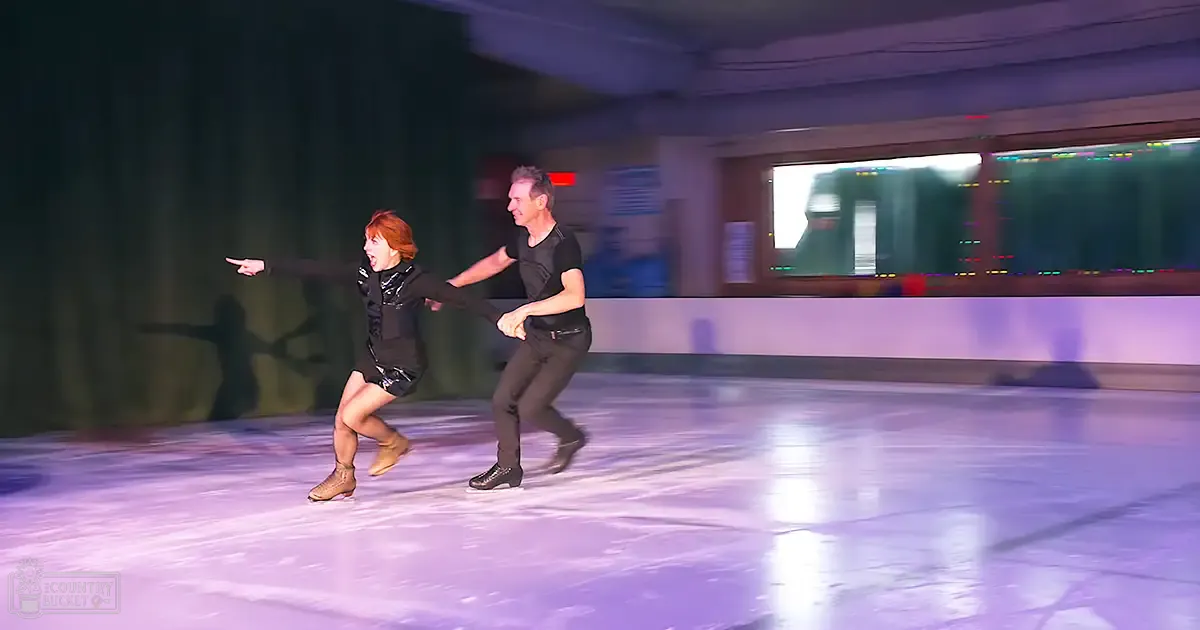 A young adult to middle-aged female skater in a black figure skating dress with a fitted bodice and flared skirt, alongside a middle-aged male skater wearing a black t-shirt and dark pants, skating at an indoor ice rink with glass barriers separating the area from spectator seating.