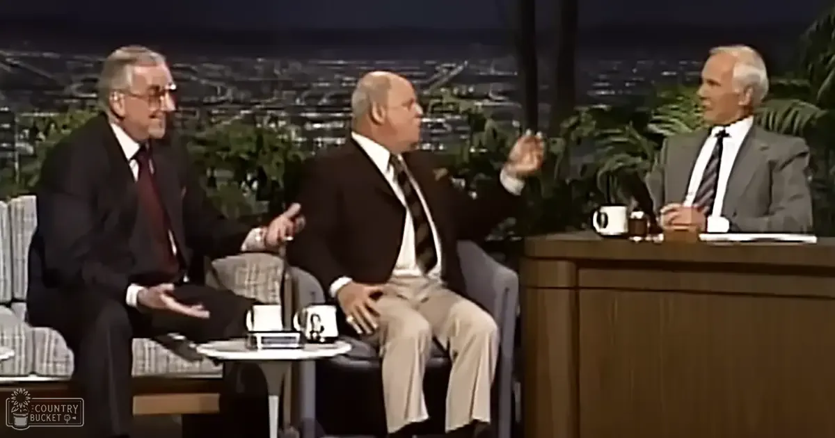Three men seated around a table on a talk show set with plants and cityscape at night.
