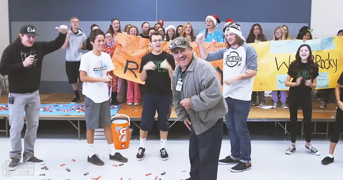 A lively indoor scene with children and adults gathered at a school event, with casual attire and an orange Gatorade cooler amidst scattered candy wrappers on the floor.