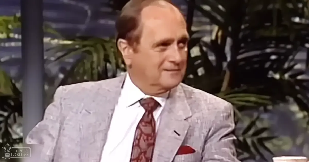 Older adult male in a light gray patterned suit, white dress shirt, red tie with leafy design, red pocket square, studio setting with plants and greenery visible in the background.