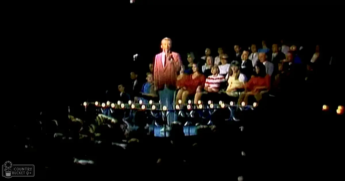 An adult male singer performing on stage in an indoor theater or concert hall; audience members seated, wearing casual attire with a red and white striped shirt prominent; nighttime setting with dim lighting