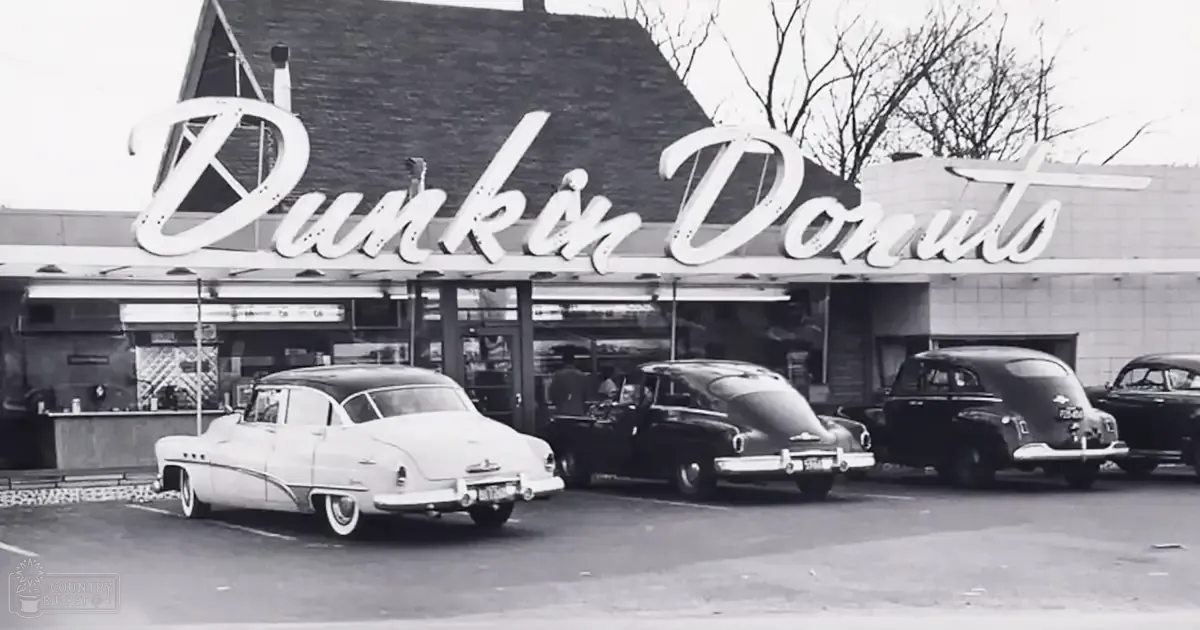 Black-and-white scene of classic cars in a parking lot at an older suburban Dunkin' Donuts location, late autumn or winter day with overcast skies and bare trees.