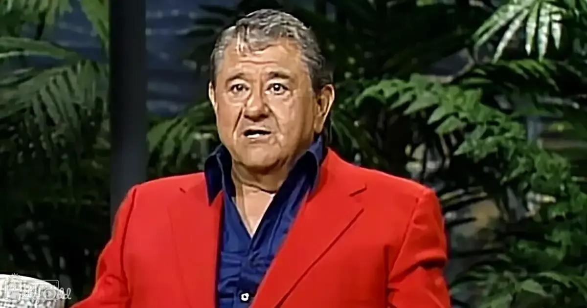 An older adult male wearing a red blazer and navy blue shirt, sitting indoors against a backdrop of green plants. Studio setting with even lighting.