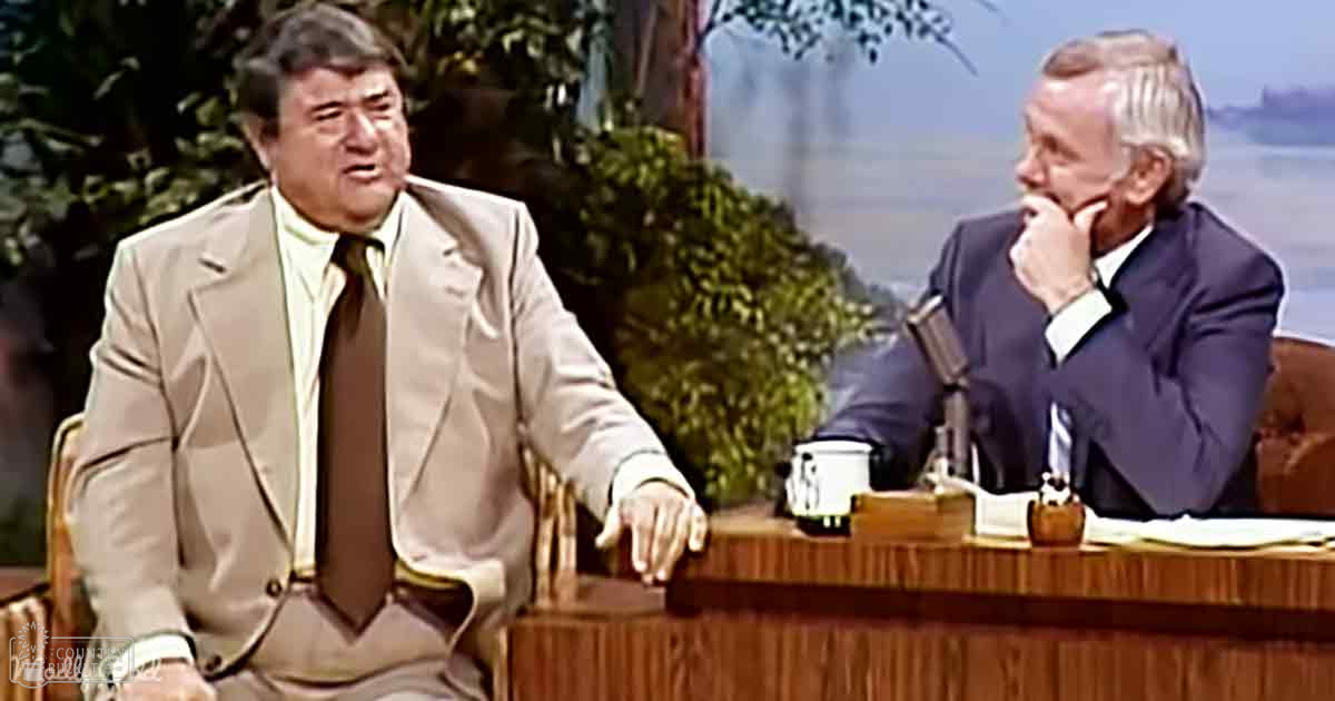 Two adults seated at a wooden desk with microphones and papers, suggesting a talk show set from the mid-20th century, framed by greenery and natural daylight.