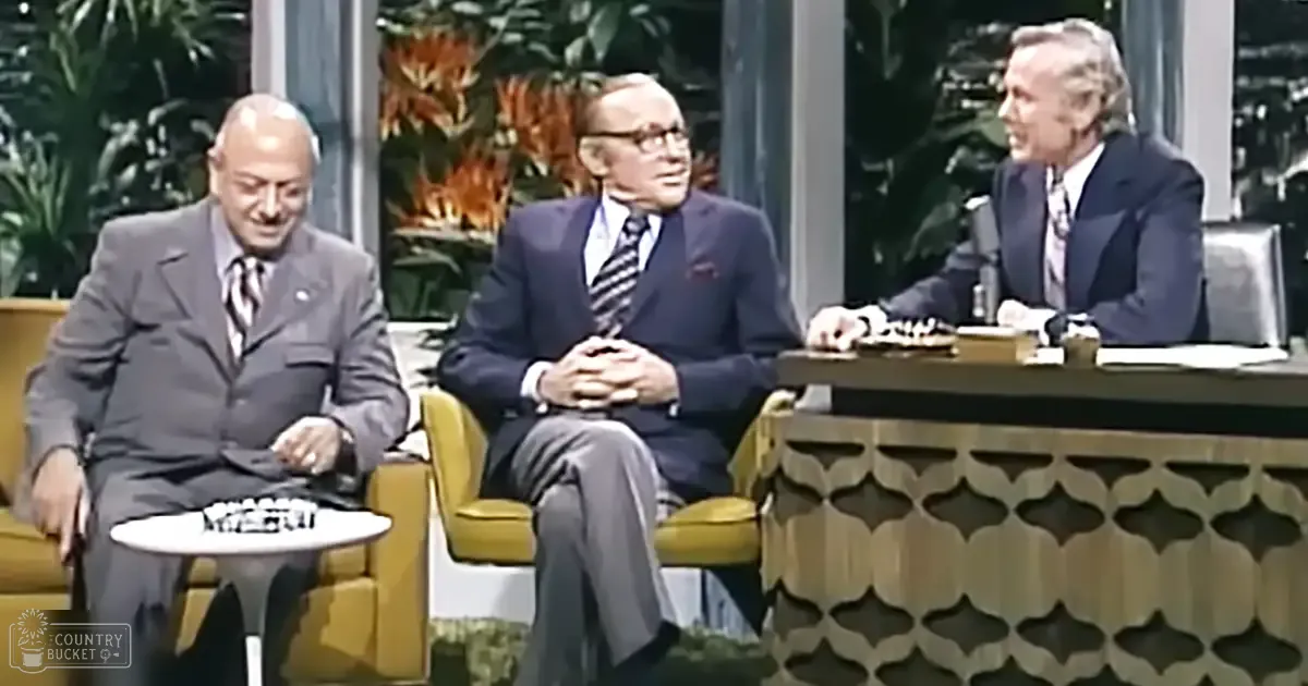 A mid-century modern studio set with three men seated on yellow armchairs, a host at the far right, large windows showing greenery outside, and natural light illuminating the scene.