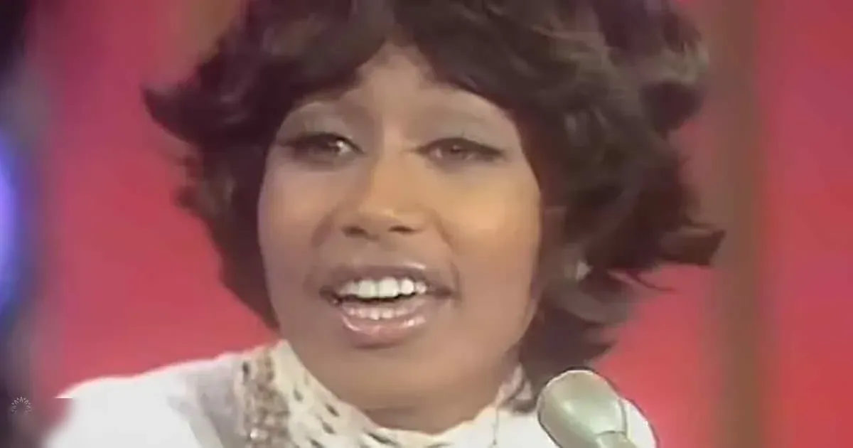 Adult female singer wearing a white patterned blouse with lace or embroidery, holding a microphone against a red backdrop in an indoor studio setting.