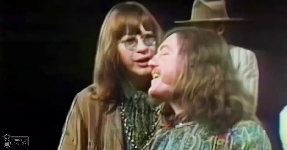 Two adults in an indoor setting; one adult female wears a long-sleeved, striped or geometric-patterned shirt and glasses; another adult male dons a floral or abstract patterned shirt with a wide-brimmed hat. Background is dark and out of focus.