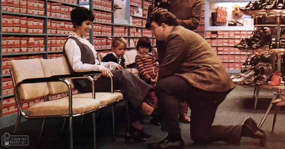 Adults assist toddlers during a shoe fitting session inside a well-lit, indoor shoe store with shelves of red shoeboxes and displays of various footwear.