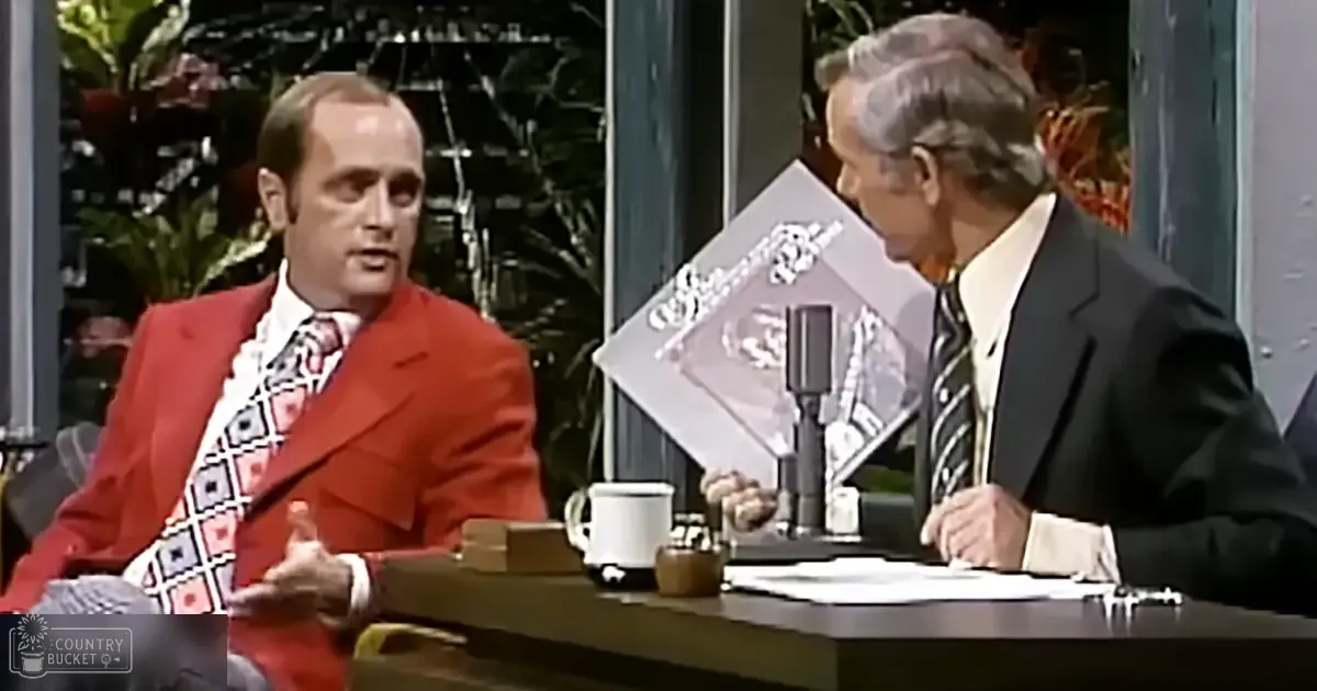 Two adults in a late 1960s to early 1970s talk show setting: one wears a bright red blazer with geometric patterned tie, the other an older adult male in a dark suit and striped tie. Decorative plants add greenery to the indoor studio background.