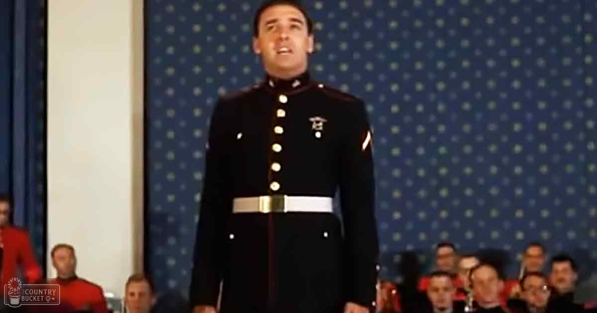 A young adult male in a black military dress uniform with white buttons and a badge stands indoors at what appears to be a formal event, possibly a military ceremony or performance. Adults in red uniforms stand behind him.