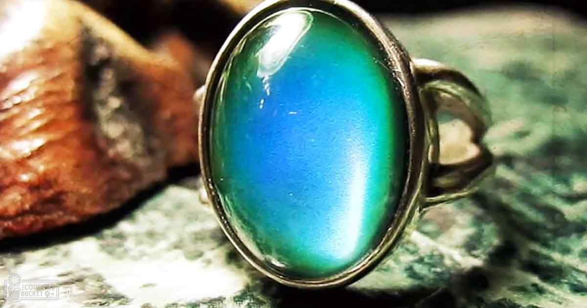 An oval-shaped gemstone ring with blue-green gradient indoors on a textured surface near a rock.
