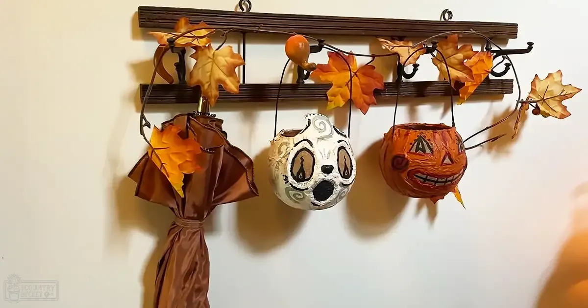 Autumn-themed indoor setting with wooden rack with hooks holding autumn leaves and decorative items; three pumpkin-shaped lanterns hanging: one white with black swirls, another orange smiling face, third brownish-orange intricate design; umbrella with orange and yellow pattern on left side of rack. Plain white background suggests wall, natural lighting indicates daytime.