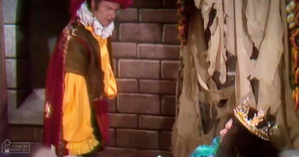 An adult male singer in a Renaissance-style yellow and maroon jacket with puffed sleeves stands beside a child wearing a blue and gold dress with long sleeves, both set against a brick wall backdrop suggesting an indoor medieval or fantasy-themed setting.