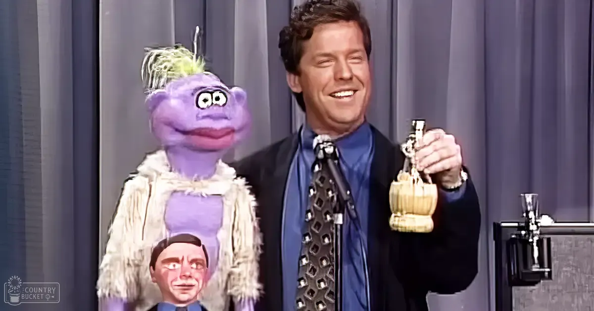 A puppeteer in a dark suit and blue shirt performs with an adult Muppet puppet on stage.