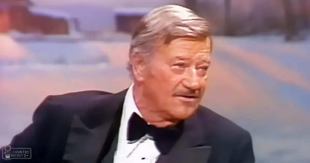 An older adult male wearing a black tuxedo with white dress shirt and black bow tie stands indoors, possibly at an event or formal gathering.