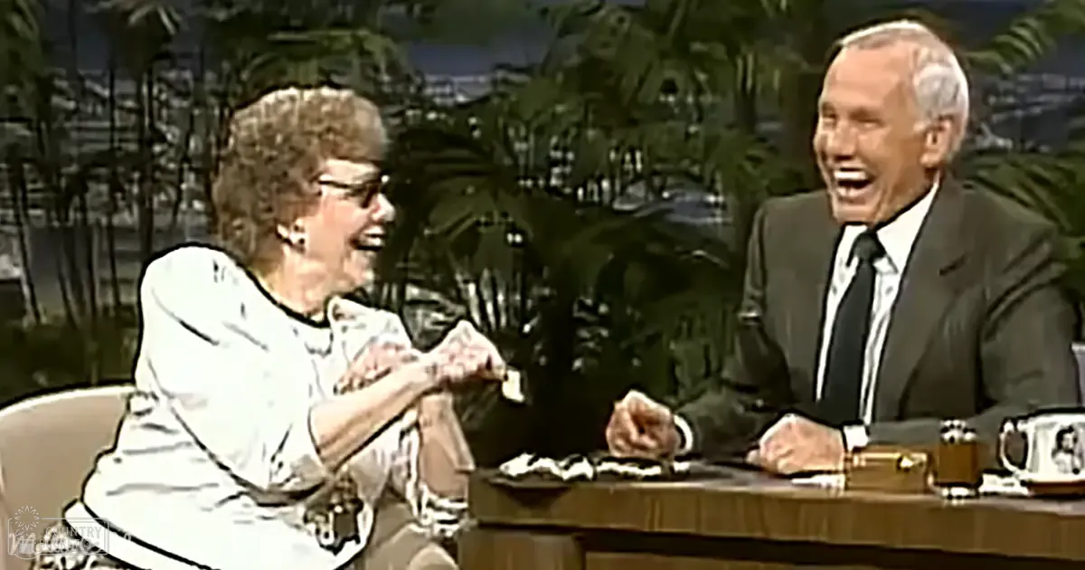 Talk show setting with two older adults seated at a desk; one in a white patterned blouse, another in a dark suit jacket and tie, both wearing formal attire typical of the mid-20th century. Decorative palm trees and tropical backdrop suggest an indoor studio designed to mimic an outdoor scene.