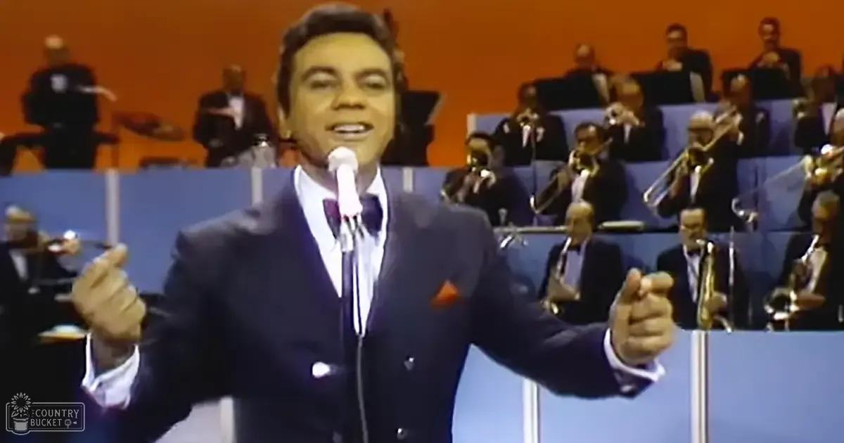 A mid-20th-century male performer stands before an orchestra, likely at a concert hall with orange walls and blue seating.