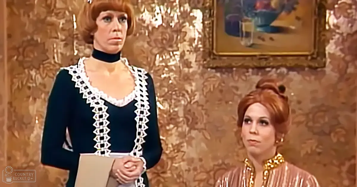 Middle-aged woman in a dark blue lace-trimmed dress stands indoors with ornate wallpaper with earthy tones; another middle-aged woman in a pinkish-gold sequined dress sits nearby, framed floral picture on the wall adds detail to the setting.