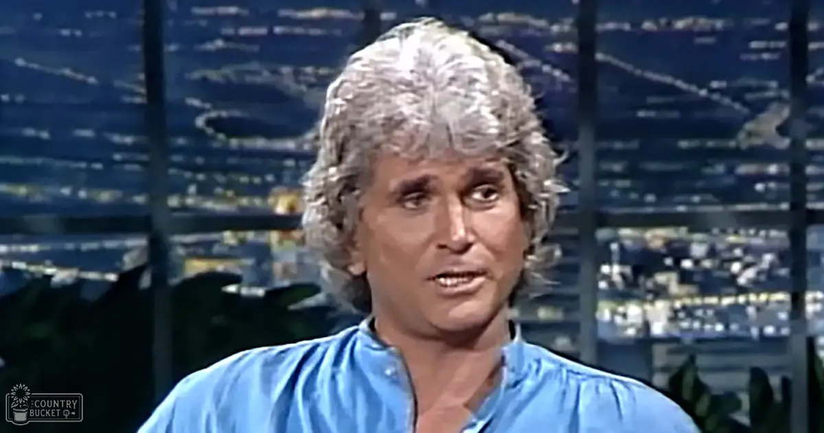 A middle-aged adult male in a relaxed, loose-fitting blue collared shirt with long sleeves stands indoors against a backdrop of large windows showing a nighttime cityscape.