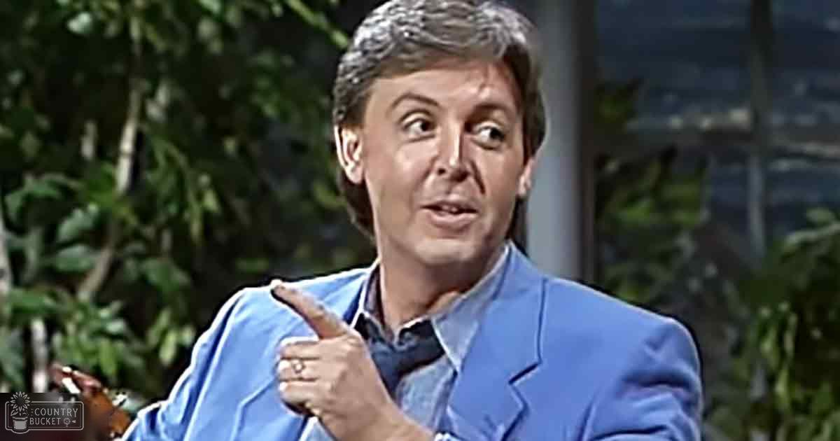 A middle-aged adult male wearing a fitted blue blazer, collared shirt, and long sleeves indoors with greenery visible through large windows in the background. The setting suggests daytime with natural light.