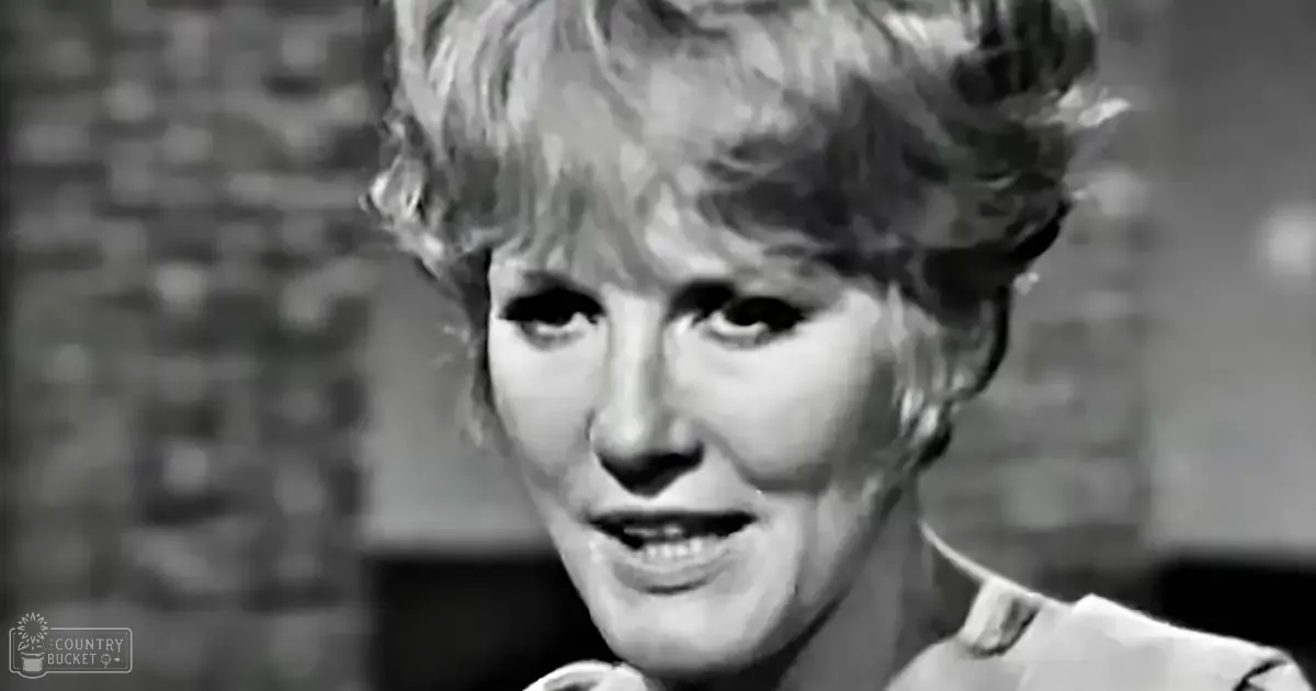 A middle-aged adult female wearing a relaxed and casual collared shirt in an indoor setting with blurred brickwork, monochrome lighting.