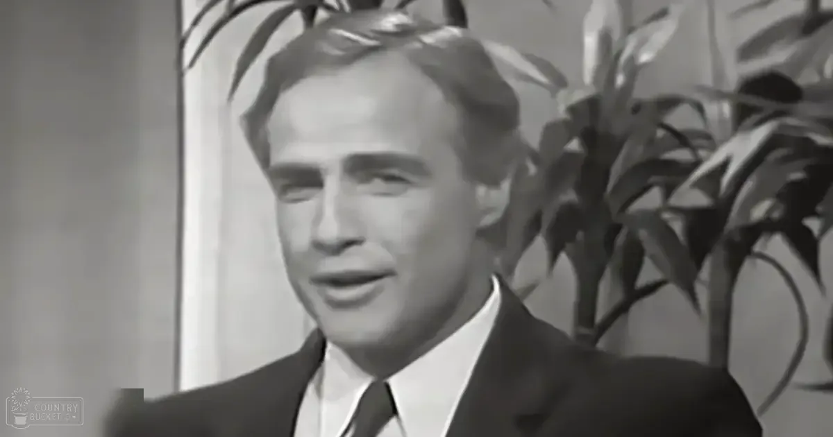 A middle-aged man wearing a dark suit jacket with crisp white shirt and dark tie in an indoor setting with soft lighting, possibly home or office environment.