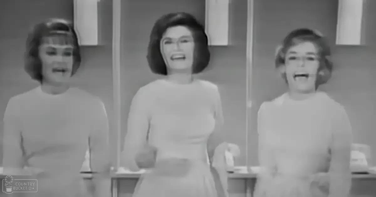 Three children, likely between 5-8 years old, dressed in mid-20th-century-style white shirts, stand together indoors with wooden cabinetry and neutral-colored walls visible behind them.
