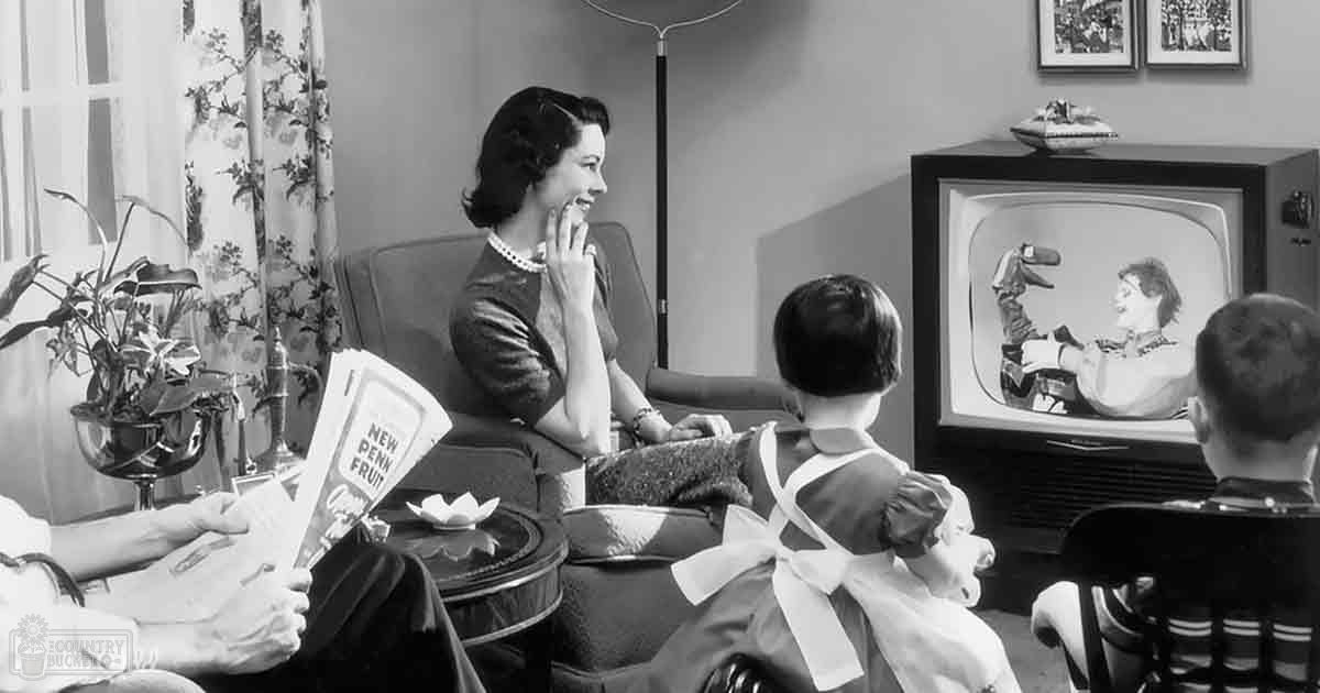 A mid-20th-century living room scene with a family dressed formally for an occasion; adult female in dark dress and pearl jewelry, child in white dress on lap, male in dark suit holding magazine titled 'New Penn Fruit'.
