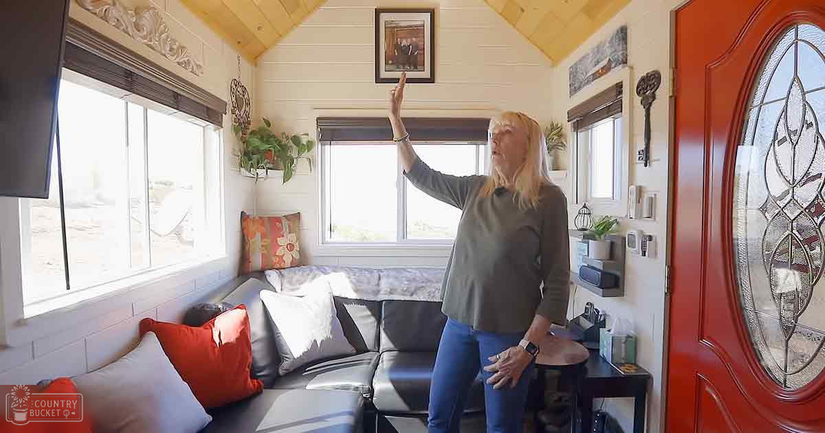 An older adult female in a small living space, possibly a tiny home. The room features furniture like a gray sectional sofa with red and white cushions, framed pictures on the walls, and decorative heart-shaped objects.