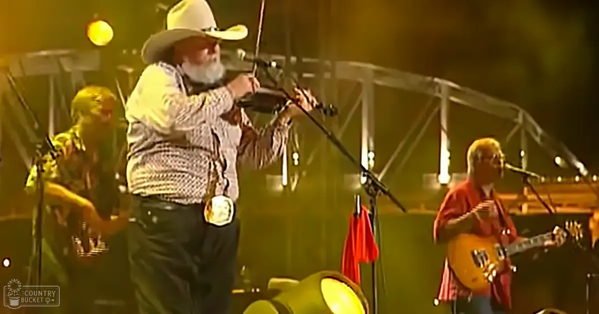 A male performer wearing a white cowboy hat, checkered shirt with small squares pattern, and dark pants plays a fiddle in an outdoor concert setting with other musicians playing electric and acoustic guitars. The stage is illuminated by yellow lights, adorned with red flags.