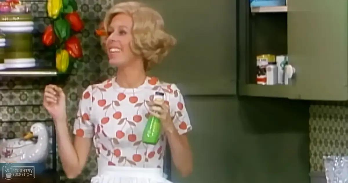 Adult female wearing a short-sleeved, above-the-knee-length white dress with red cherries and green leaves as a pattern in a retro-style kitchen setting, holding a green spray bottle.