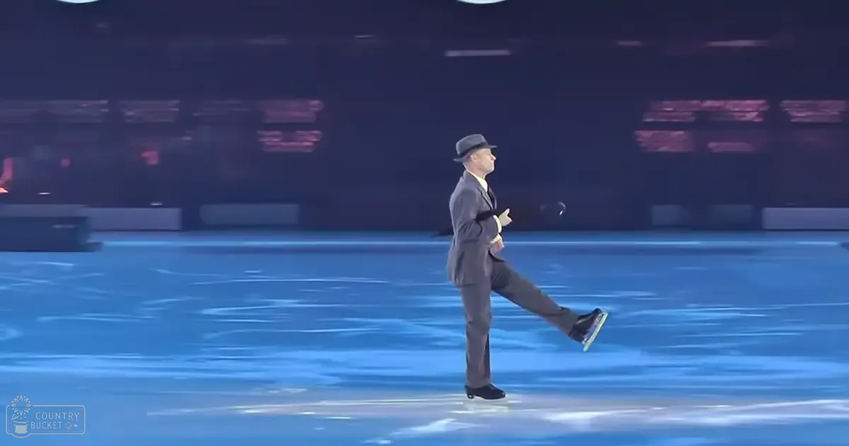 An adult male singer performing on an indoor ice rink wearing a dark gray suit, white shirt, and black fedora hat with black dress shoes, holding a small object in his right hand under dim evening lighting.