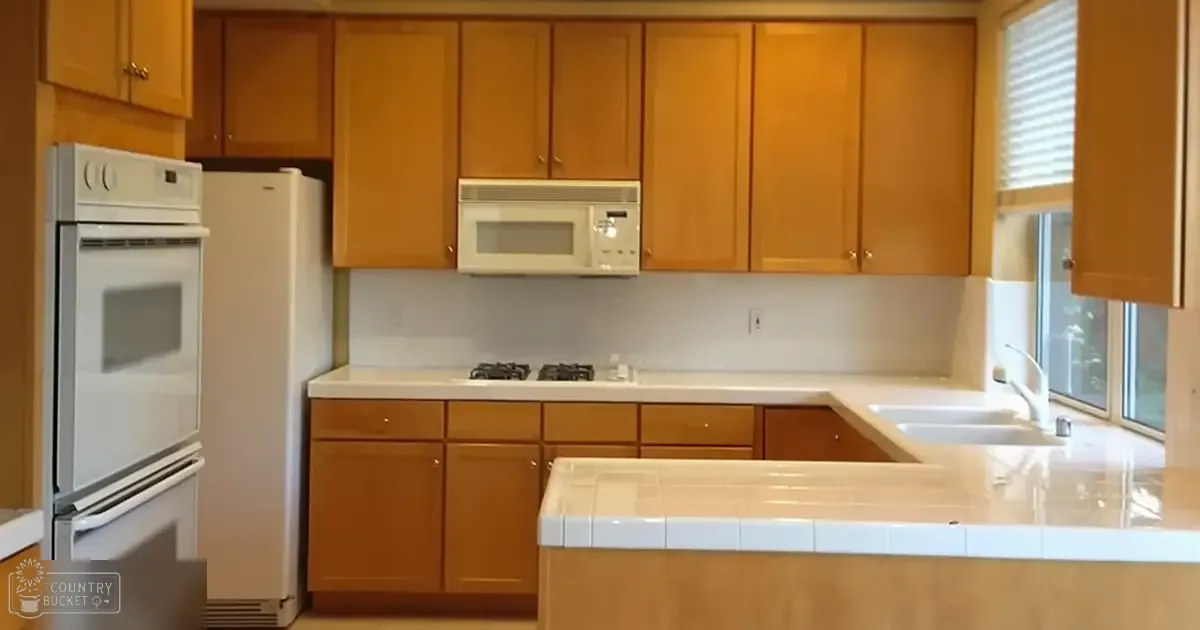 A clean, organized residential kitchen with wooden cabinets, white countertops, a double oven refrigerator, microwave above the stove, sink with faucet, window with blinds allowing natural light. The setting appears daytime due to natural light.