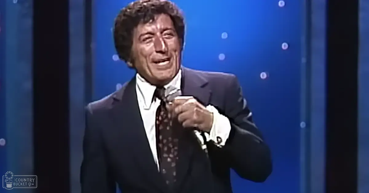 Middle-aged male performer in a dark suit, holding a microphone against a blue backdrop with circular light patterns.