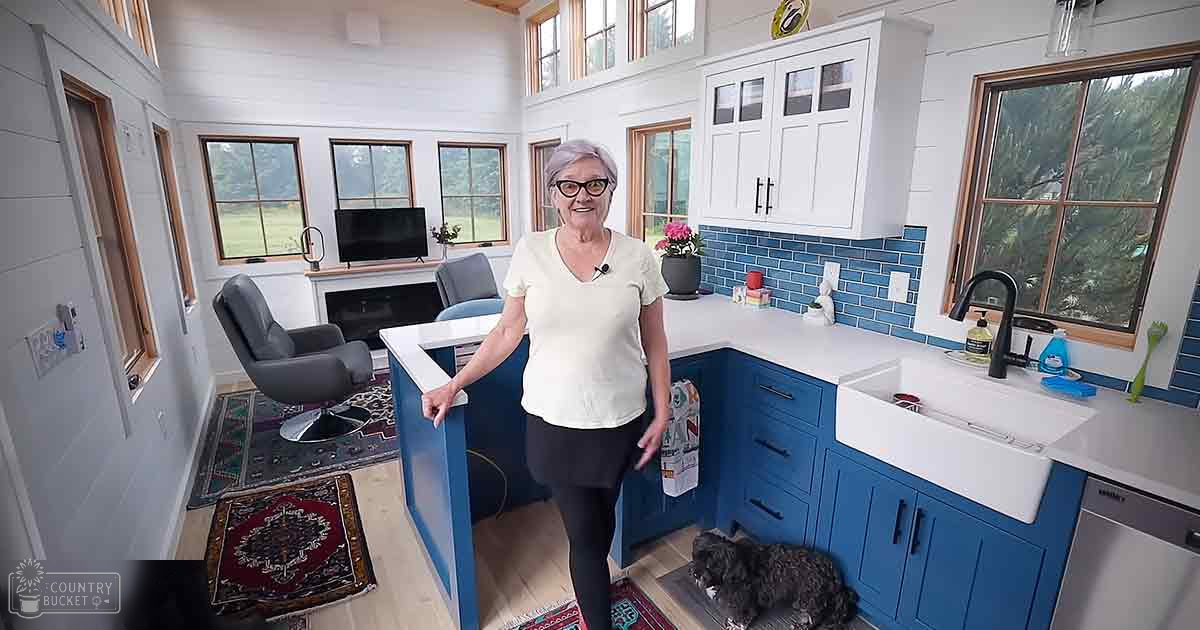 A black terrier dog lies on the floor near a kitchen counter with blue cabinets and white countertops, while an older adult female in a white t-shirt is visible in the background. The room has large windows allowing natural light into the open-concept kitchen with greenery outside.