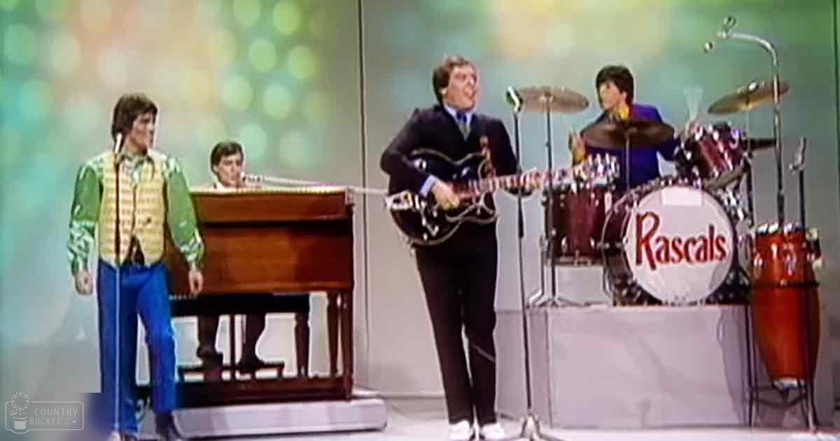 A vibrant musical performance on stage with a young adult male guitarist in a dark suit and white shoes holding an electric guitar, surrounded by other musicians including a drummer labeled 'Rascals' and a pianist. The backdrop features a bokeh effect suggesting a studio or stage environment from the 1960s or early 1970s.