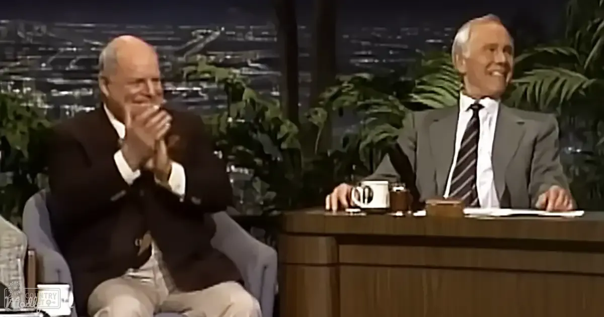 Talk show set with two older adult males in formal attire against a backdrop of palm trees and city lights at night.
