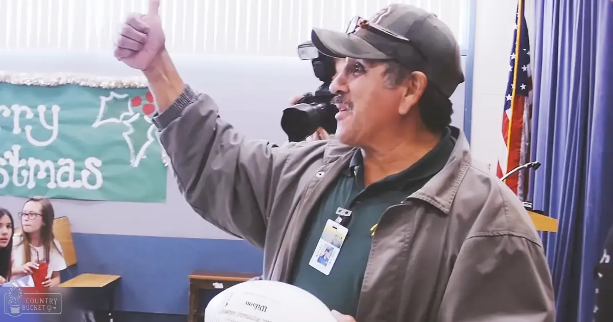 A middle-aged male giving a thumbs-up gesture in an indoor school setting with wooden furniture, a 'Merry Christmas' banner indicating holiday season, and natural daylight.