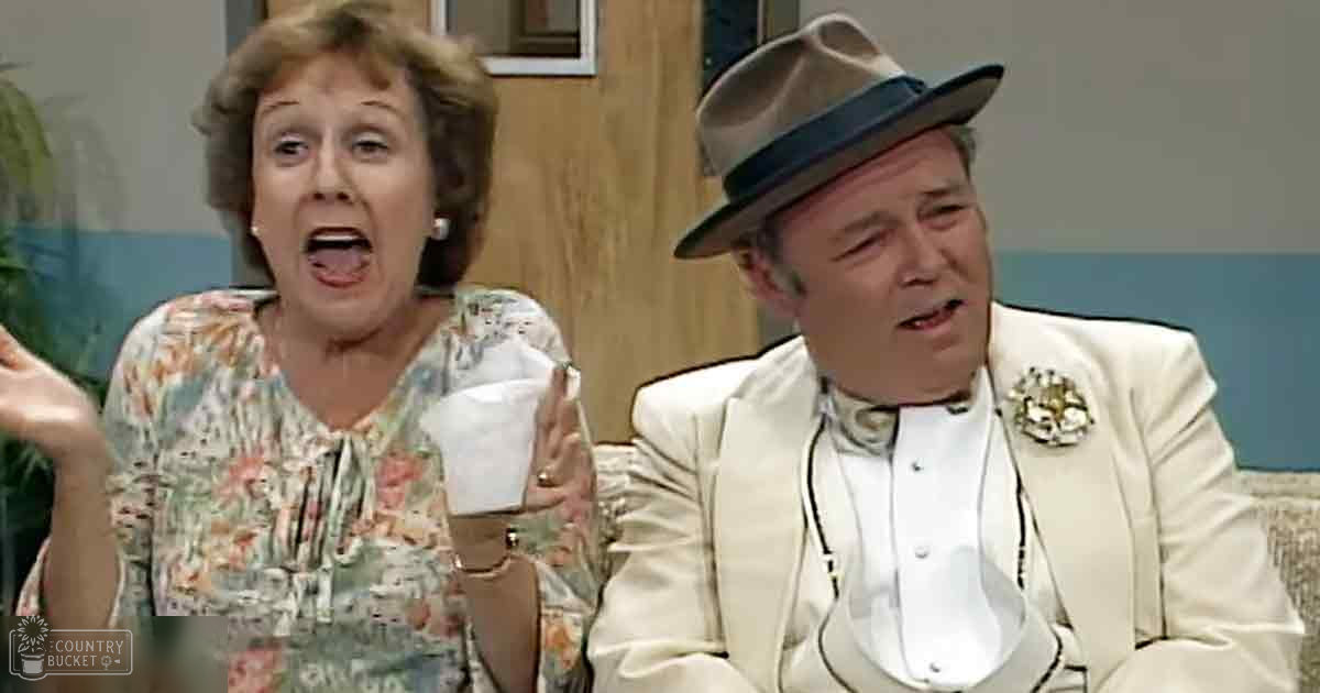 An indoor comedic scene from a television show, possibly set in a living room with two adults and domestic furnishings.