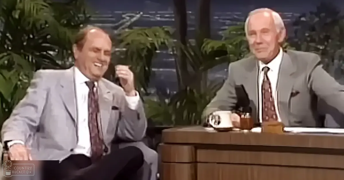 Two older adult men in a mid-20th century talk show setting with greenery and bright lighting.