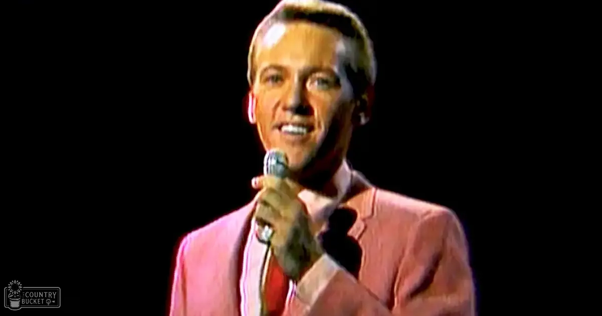 Adult male singer in a light pink blazer with vertical stripes, white shirt, and microphone on stage under professional lighting.
