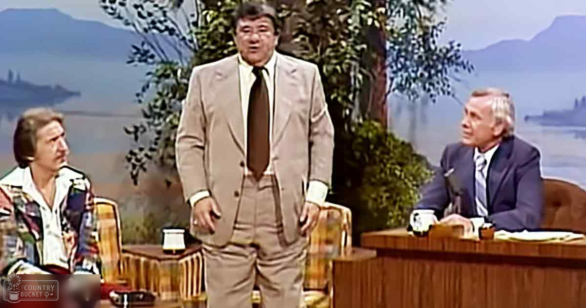 Adult male in light brown suit with white shirt and dark tie, standing behind wooden desk during a talk show set near scenic background of mountains and water.