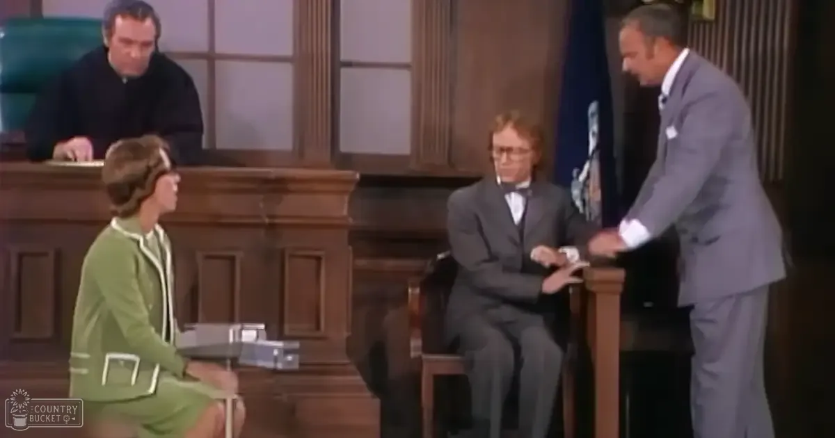 A courtroom scene with a middle-aged male in a gray suit and glasses standing at the right, an adult female in a green dress seated to the left, and another adult male in a dark suit wearing a bow tie seated center. Wooden benches and paneling suggest a formal setting.
