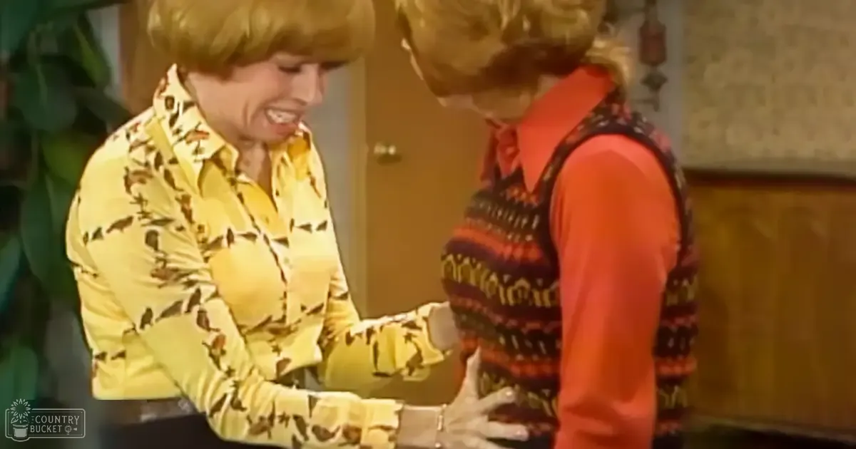 Two adults, one wearing a long-sleeved patterned yellow shirt with dark brown/black shapes from an earlier era, another in a sleeveless striped vest over an orange shirt, standing indoors under natural light.